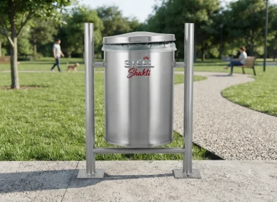 Brushed stainless steel dustbin with hinged lid, featuring the red and metallic 'STEEL Shakti' logo, securely mounted on a twin-post stand with base plates on a concrete park pathway, with a blurred park background